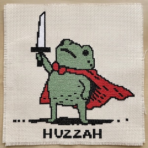 May include: A cross-stitch embroidery on beige Aida fabric featuring a pixelated green frog wearing a red cape and holding a white sword aloft. Below the frog, the word HUZZAH is stitched in black capital letters. The edges are frayed.