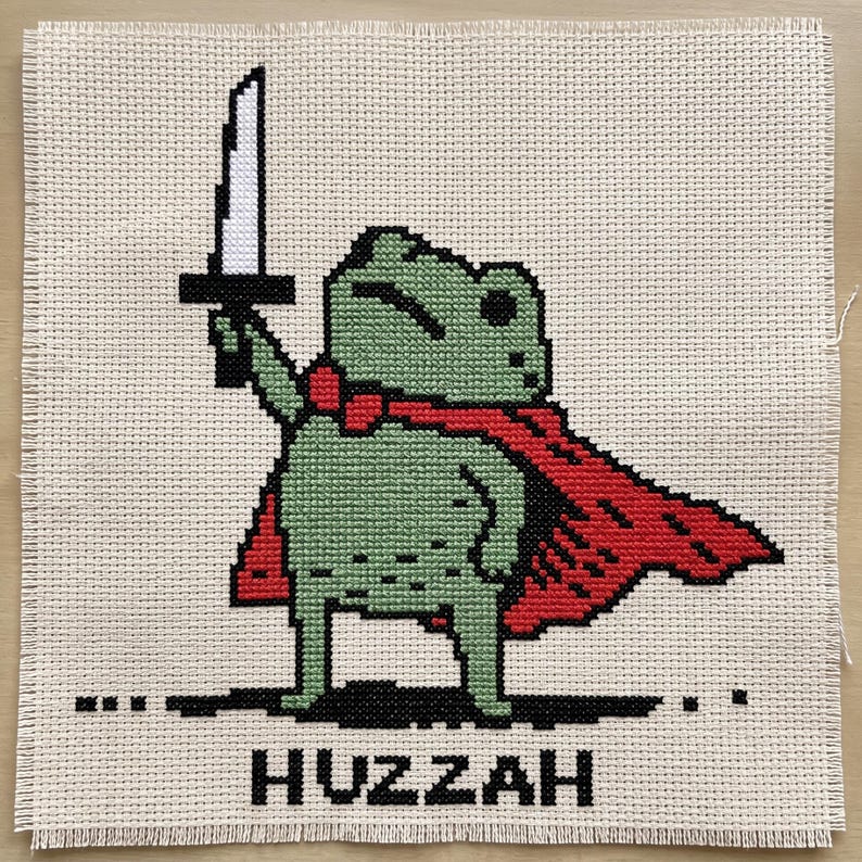 May include: A cross-stitch embroidery on beige Aida fabric featuring a pixelated green frog wearing a red cape and holding a white sword aloft. Below the frog, the word HUZZAH is stitched in black capital letters. The edges are frayed.