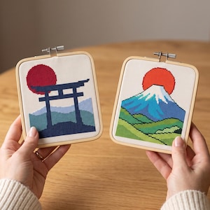Op de afbeelding: Twee vierkante houten borduurringen met kruissteekkunst. De linker ring toont een donkerblauwe torii-poort tegen een rode zon en lichtblauwe bergen. De rechter ring toont een blauwe en witte bergtop onder een rode zon.