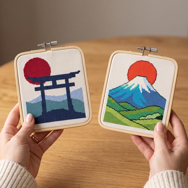 Op de afbeelding: Twee vierkante houten borduurringen met kruissteekkunst. De linker ring toont een donkerblauwe torii-poort tegen een rode zon en lichtblauwe bergen. De rechter ring toont een blauwe en witte bergtop onder een rode zon.