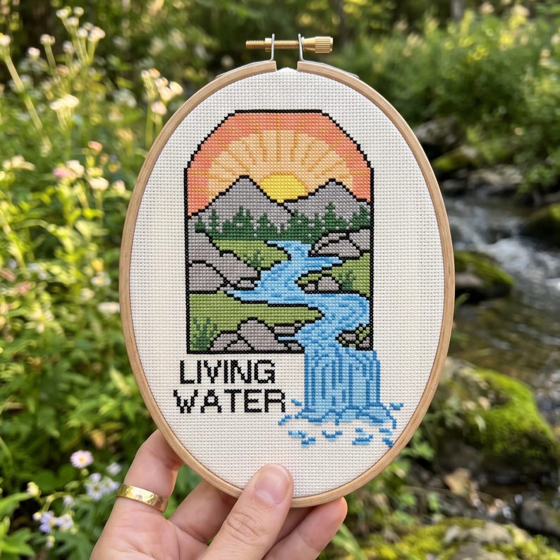 K&ouml;nnte beinhalten: Ein ovaler Stickrahmen aus Holz mit einem Kreuzstichmotiv, das eine Landschaft mit Sonnenuntergang, Bergen, B&auml;umen und einem blauen Wasserfall zeigt. Der Text "LIVING WATER" ist in schwarzen Buchstaben unten links auf dem wei&szlig;en Stoff gestickt.