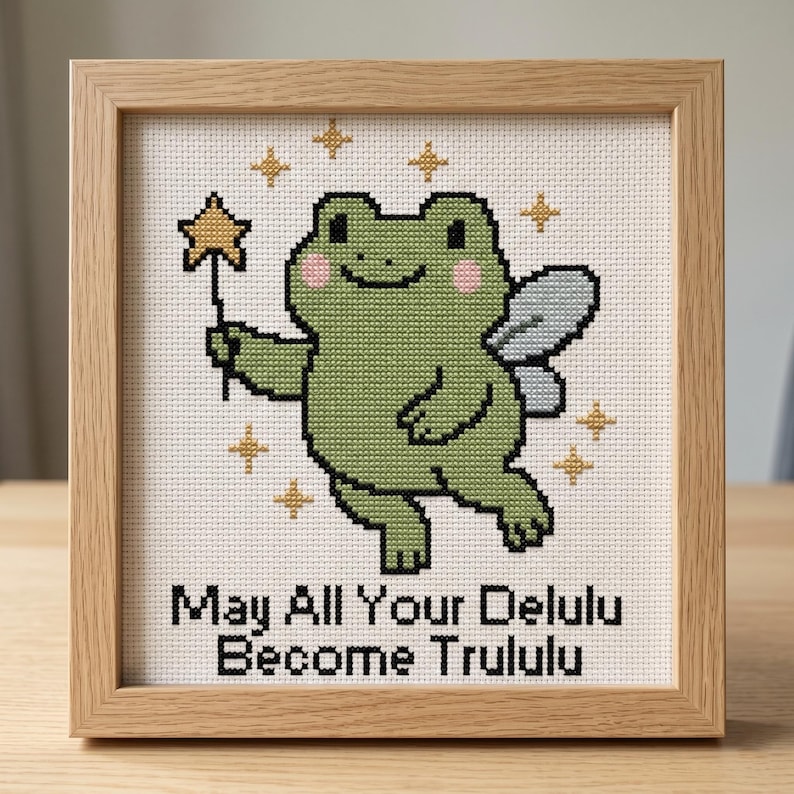 May include: Cross-stitch art in a wooden frame featuring a green frog with fairy wings and a star wand. The frog has pink cheeks and is surrounded by gold stars. Text at the bottom reads "May All Your Delulu Become Trululu."