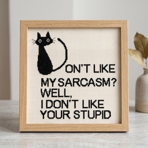 Pode incluir: Um bordado em ponto cruz quadrado numa moldura de madeira clara com a silhueta de um gato preto e o texto a preto: DON'T LIKE MY SARCASM? WELL, I DON'T LIKE YOUR STUPID. O design &eacute; bordado sobre um tecido de cor creme.