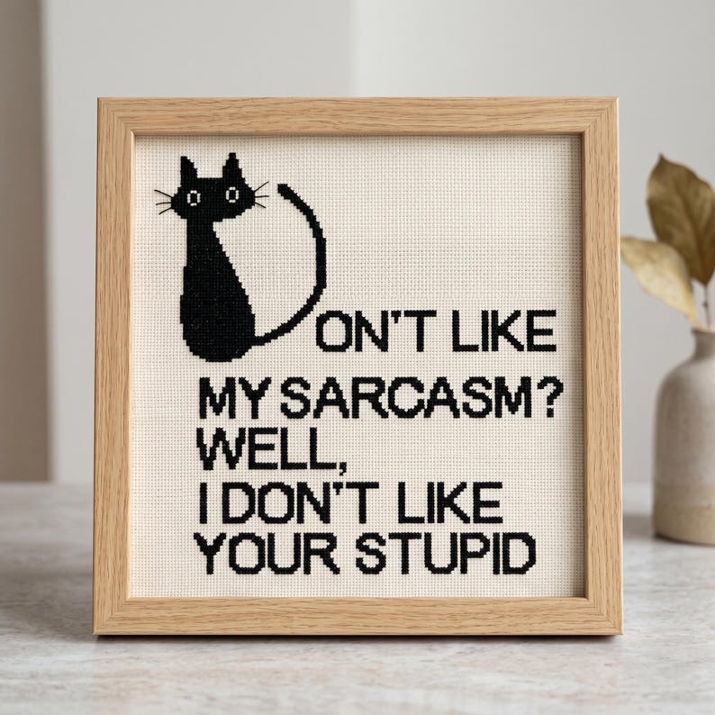 Pode incluir: Um bordado em ponto cruz quadrado numa moldura de madeira clara com a silhueta de um gato preto e o texto a preto: DON'T LIKE MY SARCASM? WELL, I DON'T LIKE YOUR STUPID. O design &eacute; bordado sobre um tecido de cor creme.