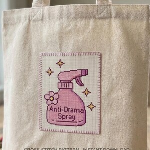 Peut inclure: Sac fourre-tout beige avec un patch en point de croix repr&eacute;sentant un vaporisateur rose. Le patch indique "Anti-Drama Spray" avec des fleurs et des &eacute;toiles. Les mots "CROSS STITCH PATTERN - INSTANT DOWNLOAD" sont en bas.