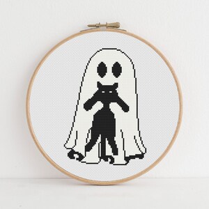 K&ouml;nnte beinhalten: Ein Kreuzstich-Stickrahmen mit einem Halloween-Design. Das Design zeigt einen wei&szlig;en Geist mit schwarzen Augen und eine schwarze Katze. Die Stickerei befindet sich in einem runden Holzrahmen.