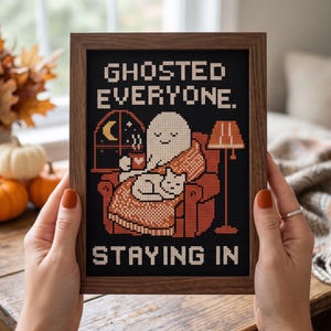 May include: A framed cross-stitch art piece featuring a white ghost sitting in an orange armchair with a cat, holding a mug. The text reads GHOSTED EVERYONE. STAYING IN. The design includes a window with a crescent moon and a floor lamp.