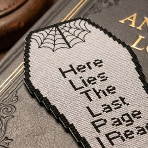 May include: A gray coffin-shaped bookmark with black text that reads "Here Lies The Last Page Read." A black spiderweb design is at the top. The bookmark has a black border and is resting on a dark book.