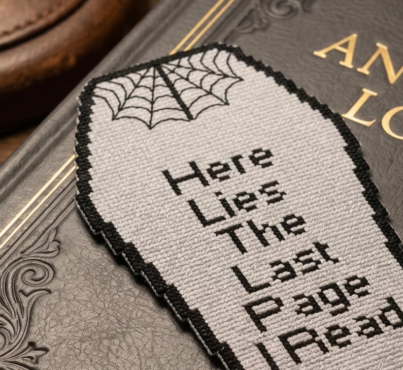May include: A gray coffin-shaped bookmark with black text that reads "Here Lies The Last Page Read." A black spiderweb design is at the top. The bookmark has a black border and is resting on a dark book.