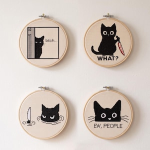 Peut inclure: Quatre tambours &agrave; broder circulaires en bois contenant des points de croix sur tissu cr&egrave;me. Chaque tambour pr&eacute;sente un chat noir avec du texte : un qui guette avec 'bitch...', un tenant un couteau rouge avec 'WHAT?', un dans l'eau et 'EW, PEOPLE'.
