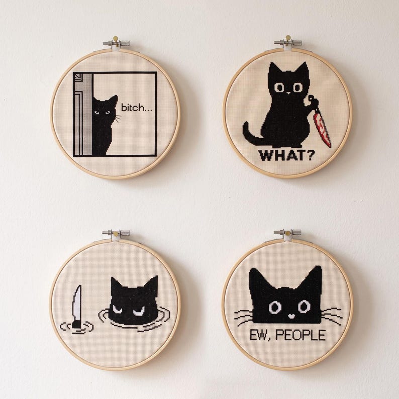 Peut inclure: Quatre tambours &agrave; broder circulaires en bois contenant des points de croix sur tissu cr&egrave;me. Chaque tambour pr&eacute;sente un chat noir avec du texte : un qui guette avec 'bitch...', un tenant un couteau rouge avec 'WHAT?', un dans l'eau et 'EW, PEOPLE'.