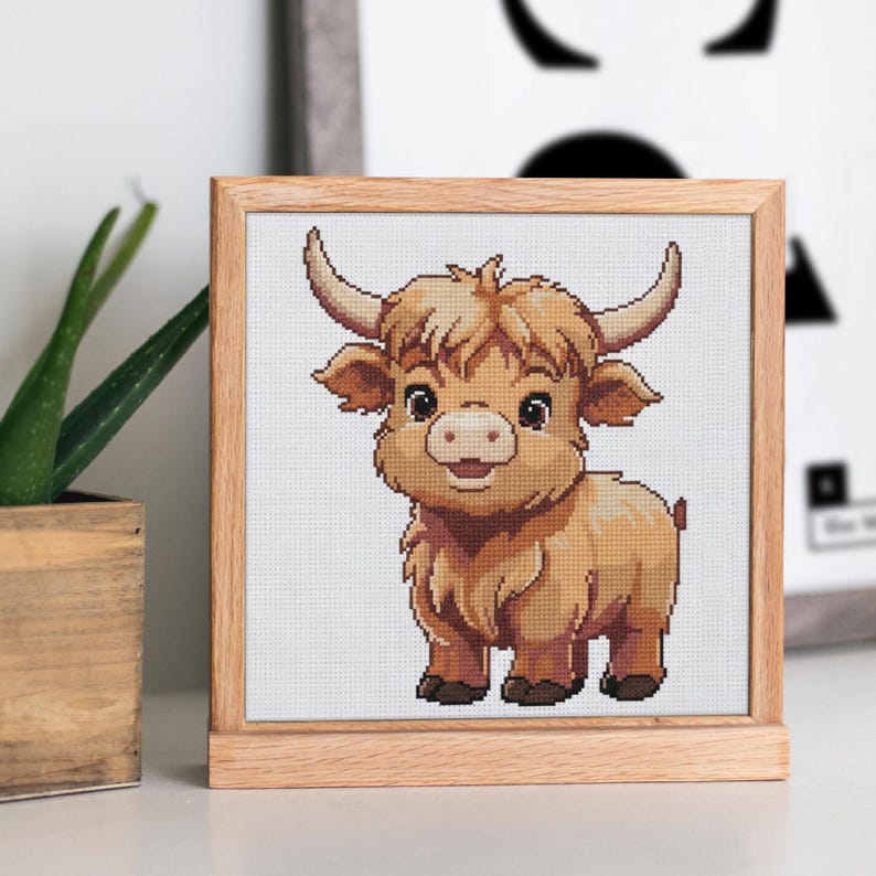 May include: A framed cross-stitch of a cartoon Highland cow. The artwork features a light brown cow with large horns, a fluffy coat, and a smiling expression. The frame is made of light-colored wood, and the background is white.