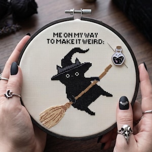 Puede incluir: Un bastidor de bordado circular con un dise&ntilde;o de punto de cruz sobre tela color crema. El dise&ntilde;o muestra un gato negro con sombrero de bruja montado en una escoba beige, con el texto ME ON MY WAY TO MAKE IT WEIRD: y un peque&ntilde;o pin de poci&oacute;n.
