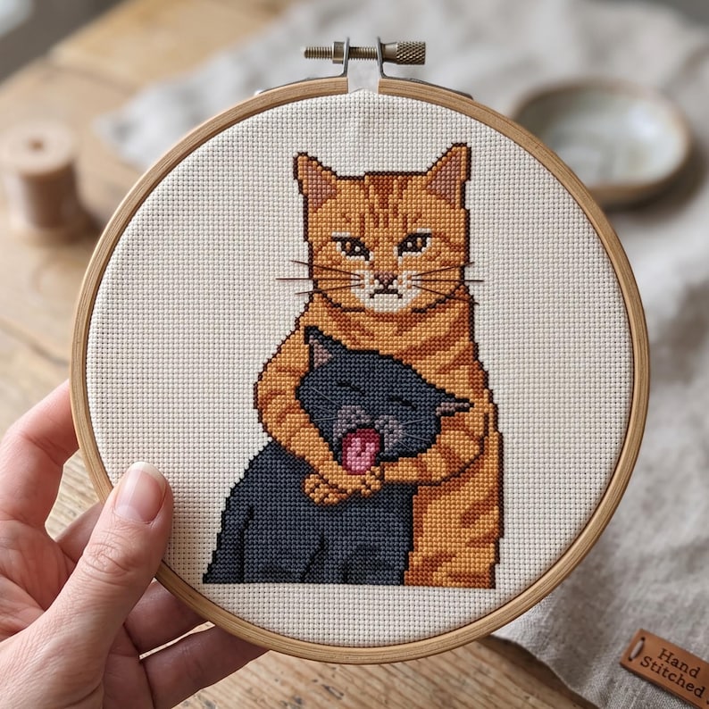 Puede incluir: Un bastidor de bordado circular de madera que contiene un dise&ntilde;o de punto de cruz de un gato atigrado naranja abrazando a un gato m&aacute;s peque&ntilde;o de color gris oscuro. El gato naranja tiene una expresi&oacute;n seria y el gris tiene la boca abierta.