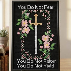 May include: A framed cross stitch pattern on a black background featuring a central sword surrounded by pink flowers and green leaves. The text reads: You Do Not Fear, You Do Not Falter, You Do Not Yield. The design is presented in a black frame.