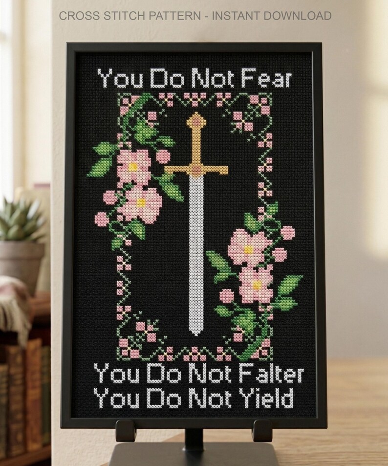 May include: A framed cross stitch pattern on a black background featuring a central sword surrounded by pink flowers and green leaves. The text reads: You Do Not Fear, You Do Not Falter, You Do Not Yield. The design is presented in a black frame.