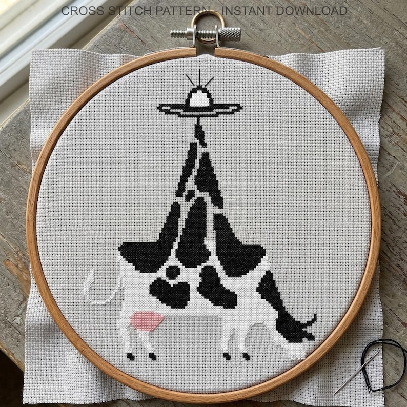 Pode incluir: Um padr&atilde;o de ponto cruz de uma vaca com manchas pretas e brancas e um OVNI acima. O design est&aacute; num bastidor de bordado de madeira, com o texto "CROSS STITCH PATTERN - INSTANT DOWNLOAD" no topo. Uma agulha e linha s&atilde;o vis&iacute;veis.
