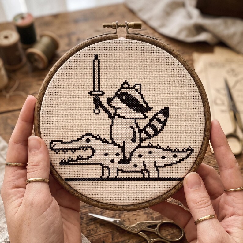 May include: A circular wooden embroidery hoop containing a piece of cream-colored fabric with a black cross-stitch design. The pattern depicts a raccoon holding a sword while riding on the back of a crocodile, rendered in a pixelated style.