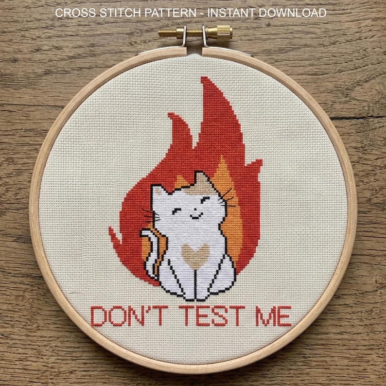 Pu&ograve; includere: Ricamo a punto croce in un telaio di legno. Il disegno presenta un gatto bianco con accenti arancioni e marroni, su uno sfondo di fiamme rosse. Il testo "DON'T TEST ME" &egrave; ricamato sotto il gatto. Il testo in alto recita "CROSS STITCH PATTERN - INSTANT DOWNLOAD".