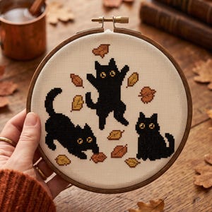 May include: A circular wooden embroidery hoop containing a piece of cream-colored fabric with a cross-stitch design of three black cats surrounded by scattered orange, yellow, and brown autumn leaves. The cats have large, wide, yellow eyes.