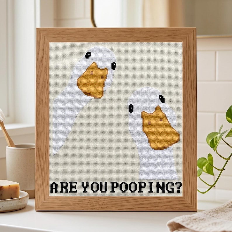 May include: A framed cross-stitch embroidery featuring two white geese with yellow beaks against a light beige background. The text ARE YOU POOPING? is stitched in black capital letters at the bottom of the piece, which is encased in a light wood frame.