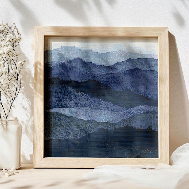 May include: A framed cross-stitch landscape depicting a mountain range in various shades of blue. The artwork is presented in a light-coloured wooden frame. The cross-stitch technique gives a pixelated appearance, adding texture to the image.