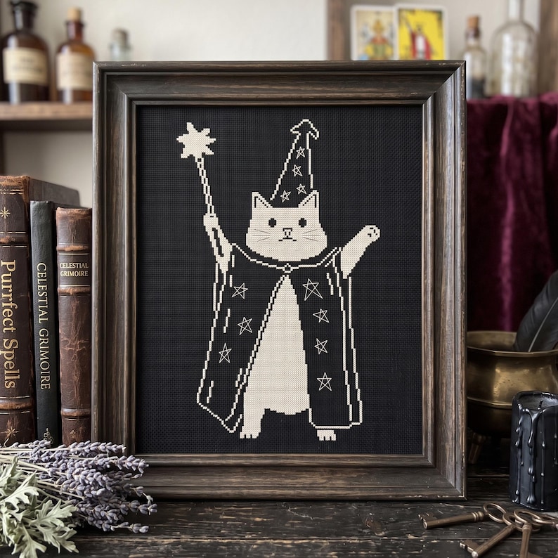 May include: A framed cross-stitch embroidery featuring a white cat dressed as a wizard in a black robe and pointed hat, both decorated with white star outlines. The cat holds a star-tipped wand in one paw, set against a solid black background.