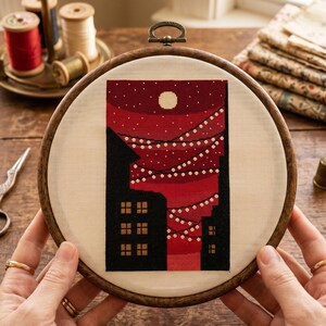 K&ouml;nnte beinhalten: Ein runder Stickrahmen aus Holz mit einem Kreuzstichmotiv auf cremefarbenem Stoff. Das Design zeigt einen dunkelroten Nachthimmel mit einem wei&szlig;en Mond und h&auml;ngenden Lichterketten &uuml;ber schwarzen Geb&auml;udesilhouetten mit kleinen gelben Fenstern.