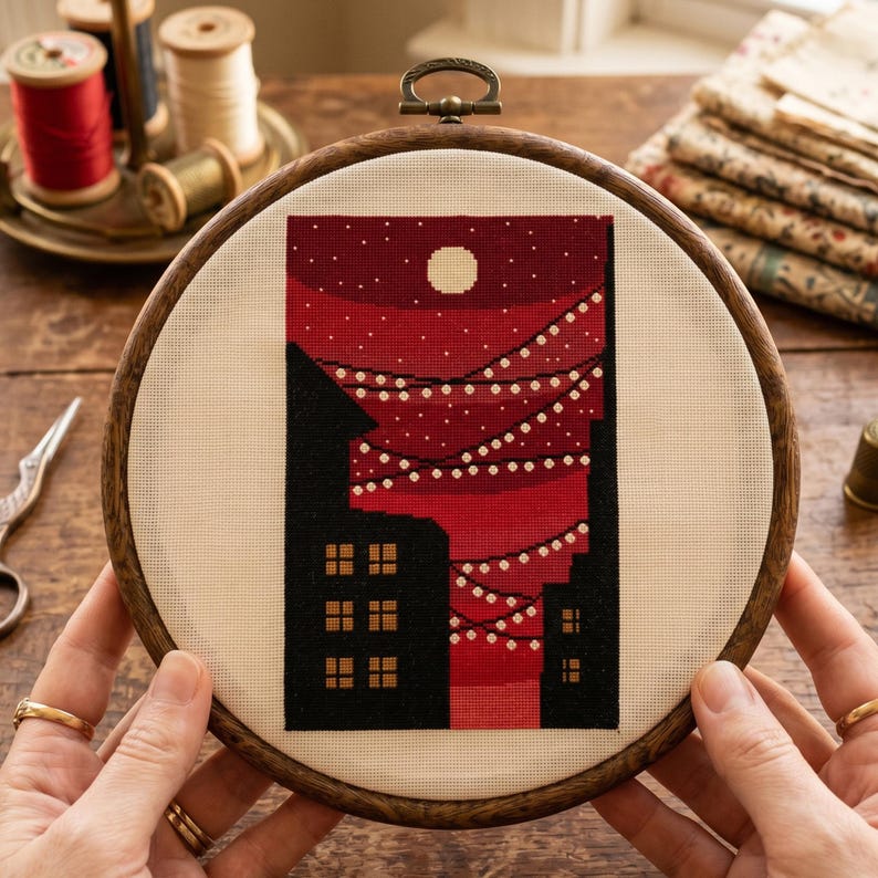 K&ouml;nnte beinhalten: Ein runder Stickrahmen aus Holz mit einem Kreuzstichmotiv auf cremefarbenem Stoff. Das Design zeigt einen dunkelroten Nachthimmel mit einem wei&szlig;en Mond und h&auml;ngenden Lichterketten &uuml;ber schwarzen Geb&auml;udesilhouetten mit kleinen gelben Fenstern.