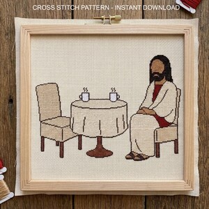 May include: Cross-stitch pattern featuring a religious figure seated at a table with two cups of coffee. The design includes a table, two chairs, and the text "CROSS STITCH PATTERN - INSTANT DOWNLOAD" at the top. The artwork is framed in a wooden hoop.