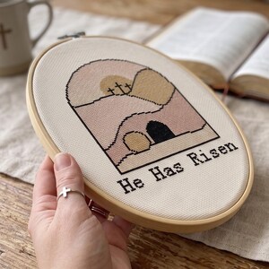 K&ouml;nnte beinhalten: Ein runder Stickrahmen aus Holz mit cremefarbenem Stoff und einem Kreuzstichmotiv. Das Design zeigt ein Grab, drei Kreuze auf einem H&uuml;gel und den in schwarzem Garn gestickten Text "He Has Risen" in verschiedenen Rosa- und Beiget&ouml;nen.
