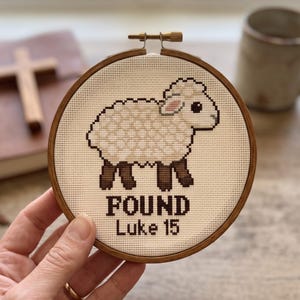 K&ouml;nnte beinhalten: Ein runder Holzstickrahmen mit einem Kreuzstichmotiv eines cremefarbenen Schafs mit dunkelbraunen Beinen und Ohren. Unter dem Schaf steht der Text "FOUND" in fetten Buchstaben und "Luke 15" in einer kleineren Schrift auf cremefarbenem Stoff.