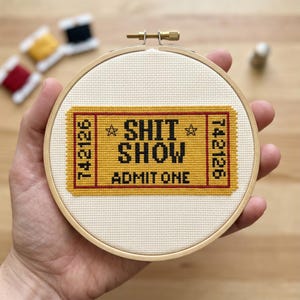 May include: A circular wooden embroidery hoop containing a cross-stitch design on off-white fabric. The design features a yellow ticket with a red border, black text reading SHIT SHOW and ADMIT ONE, and the number 742126 on both sides.