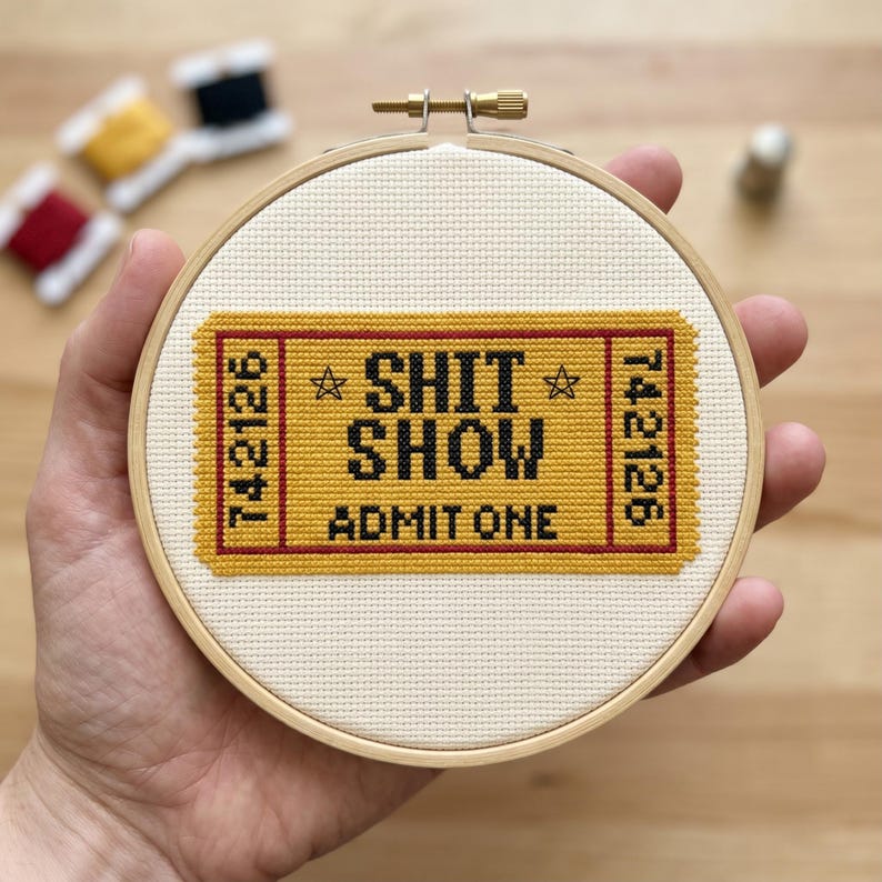 K&ouml;nnte beinhalten: Ein runder Stickrahmen aus Holz mit einem Kreuzstichmotiv auf cremefarbenem Stoff. Das Design zeigt ein gelbes Ticket mit rotem Rand, schwarzem Text mit der Aufschrift SHIT SHOW und ADMIT ONE sowie der Nummer 742126 auf beiden Seiten.