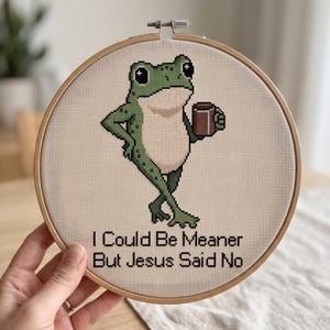 May include: A circular wooden embroidery hoop containing a cross-stitch design of a green frog holding a brown mug. The frog stands with crossed legs. Below the frog, the text reads: I Could Be Meaner But Jesus Said No in black letters.