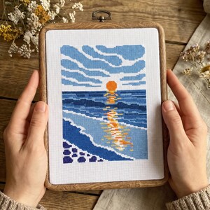 Pu&ograve; includere: Un telaio da ricamo rettangolare in legno contenente un pezzo a punto croce che raffigura un tramonto sull'oceano. Il design presenta un sole arancione brillante, onde blu e un cielo azzurro con nuvole, il tutto ricamato su tessuto bianco.