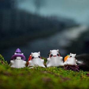 May include: A whimsical Halloween-themed display featuring four small, white ghost figurines with black eyes and mouths. One ghost wears a purple witch hat. A small orange pumpkin is also present. The figurines are set on green moss.