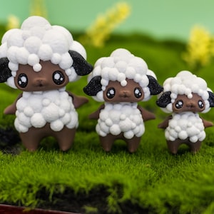 Könnte beinhalten: Drei Schaf-Figuren mit weißer, flauschiger Wolle, braunen Körpern und schwarzen Ohren stehen auf grünem Moos. Die Figuren sind unterschiedlich groß, die größte in der Mitte. Die Schafe haben große, ausdrucksstarke Augen.