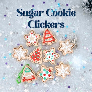 May include: A selection of sugar cookie clickers in star and Christmas tree designs. The cookies are decorated with white, red, and green icing. The words "Sugar Cookie Clickers" are in navy blue script above the cookies. A silver keyring is attached to one cookie.