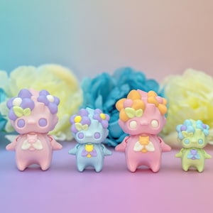 Puede incluir: Cuatro figuritas de color pastel con coronas de flores. Las figuritas son rosas, azules, verdes y rosas. Están dispuestas en fila sobre una superficie rosa claro.