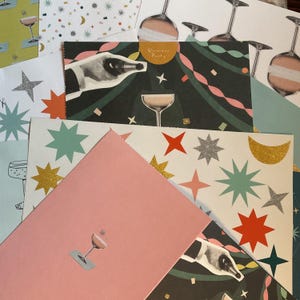 Puede incluir: Una colección de tarjetas de felicitación con diseños de champán y estrellas. Las tarjetas presentan varios patrones, incluyendo estrellas, lunas y botellas de champán. Una tarjeta tiene el texto "Glamour Party". La paleta de colores incluye rosa, verde, dorado y blanco.