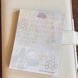 May include: A beige planner with a white sticker featuring a bee, honeycomb pattern, and the text "@sandrad_designco" and "@buzybeeplannergirl".