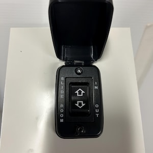 May include: A black control panel with a hinged cover. The panel has a rocker switch with up and down arrows, and the words "IN," "OUT," and "SLIDE ROOM" are printed on the panel. The cover is open.