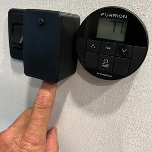 May include: A black Furrion air conditioner control panel with a digital display showing 77 degrees Fahrenheit. The control panel has buttons for fan, mode, and up and down arrows to adjust the temperature.