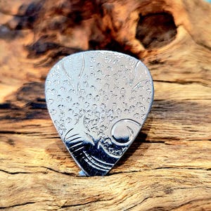 May include: A silver guitar plectrum with an engraved design. The plectrum has a teardrop shape and features a detailed pattern of leaves and swirls. The plectrum is displayed on a wooden surface.