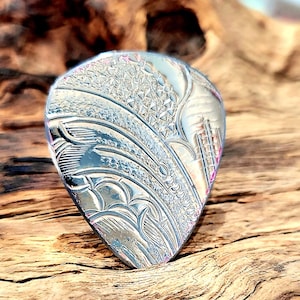 May include: A silver guitar plectrum with detailed engraved patterns. The plectrum has a teardrop shape and features intricate designs, including floral and geometric elements. The metallic surface reflects light. A musical accessory.