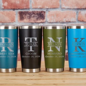 Puede incluir: Cuatro vasos personalizados de acero inoxidable en verde azulado, negro, verde oliva y azul. Cada vaso presenta un nombre y un título, con la fecha "28 de octubre de 2024" grabada debajo. Los vasos están sobre una superficie de madera.