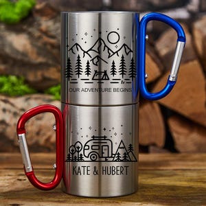 May include: Two stainless steel camping mugs stacked together. The top mug has a blue carabiner handle and features a mountain and forest design with the text "OUR ADVENTURE BEGINS." The bottom mug has a red carabiner handle and a camper design with the text "KATE & HUBERT."