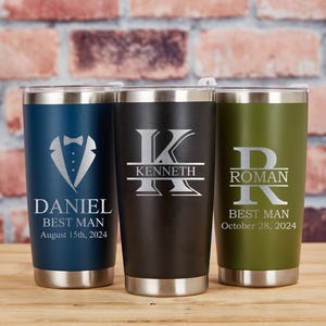 May include: Three personalized stainless steel tumblers in navy blue, black, and olive green. Each tumbler features custom text and a design, including a tuxedo graphic and initials. The tumblers are designed for a wedding party.