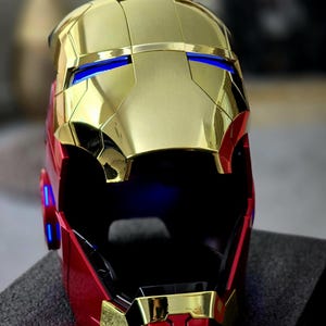 May include: A gold and red Iron Man helmet with a black visor and blue lights. The helmet is made of plastic and has a detailed design.